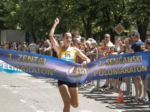 Női győztes szakítja át a kétnyelvű célszalagot, ököl a magasban, mögötte szurkolók